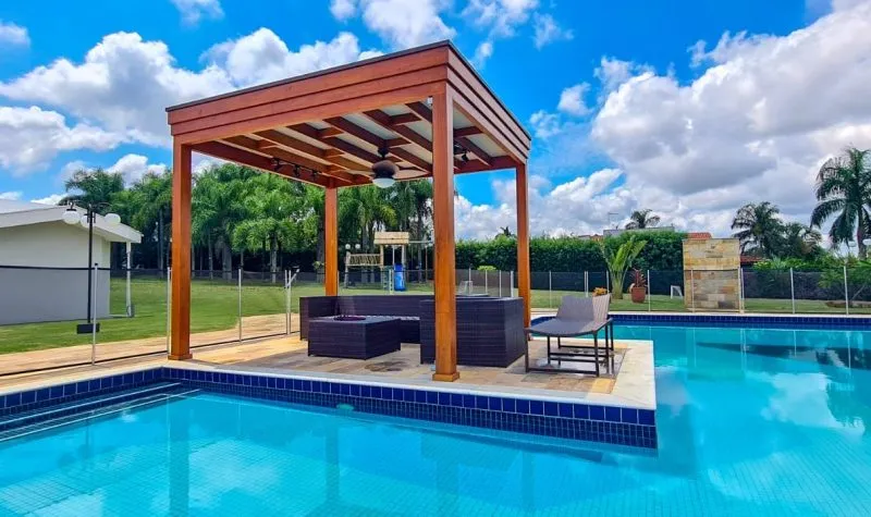 Pergolado de madeira sobre a borda da piscina, com sofás de área externa e ventilador de teto